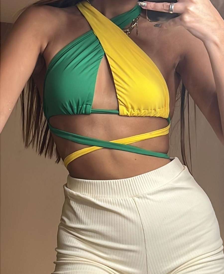 Imagem com fundo de parede bege. No centro, mulher usando roupas Brazilcore: top verde e amarelo com amarrações + short braco.