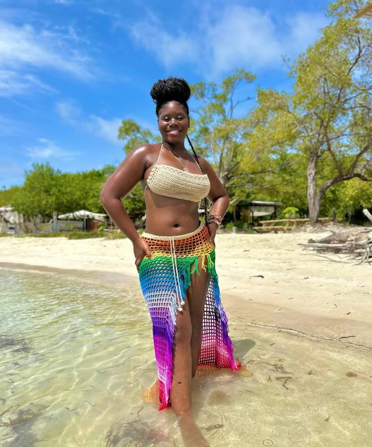 Uma mulher negra, sorridente, com coque no cabelo, posa na beira da água em uma praia tropical. Ela usa um top de biquíni de crochê bege e uma saia longa de crochê com franjas, em degradê de cores do arco-íris.
