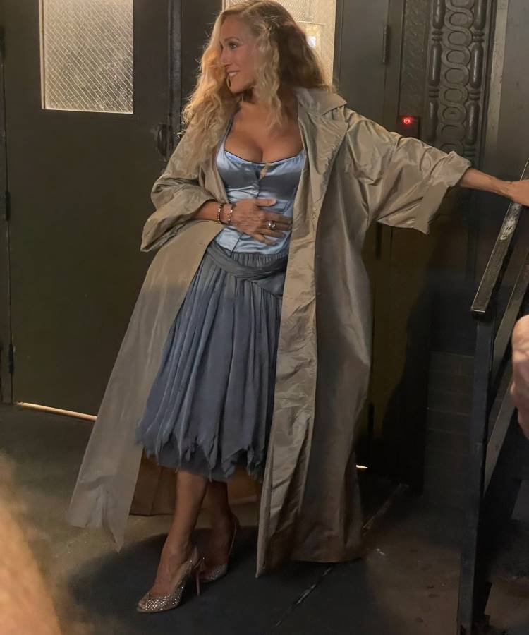 Uma mulher loira com cabelos longos e ondulados, usando um casaco comprido cor de areia sobre um vestido azul-acinzentado, incluindo um corpete e uma saia irregular. Ela usa sapatos de salto alto brilhantes e está inclinada em uma estrutura escura, com um fundo de porta metálica.
