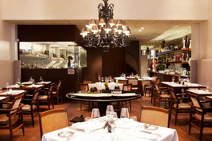 Área com cozinha de vidro, lustre e mesas de madeira. Restaurante D.O.M. - Restaurantes icônicos em SP
