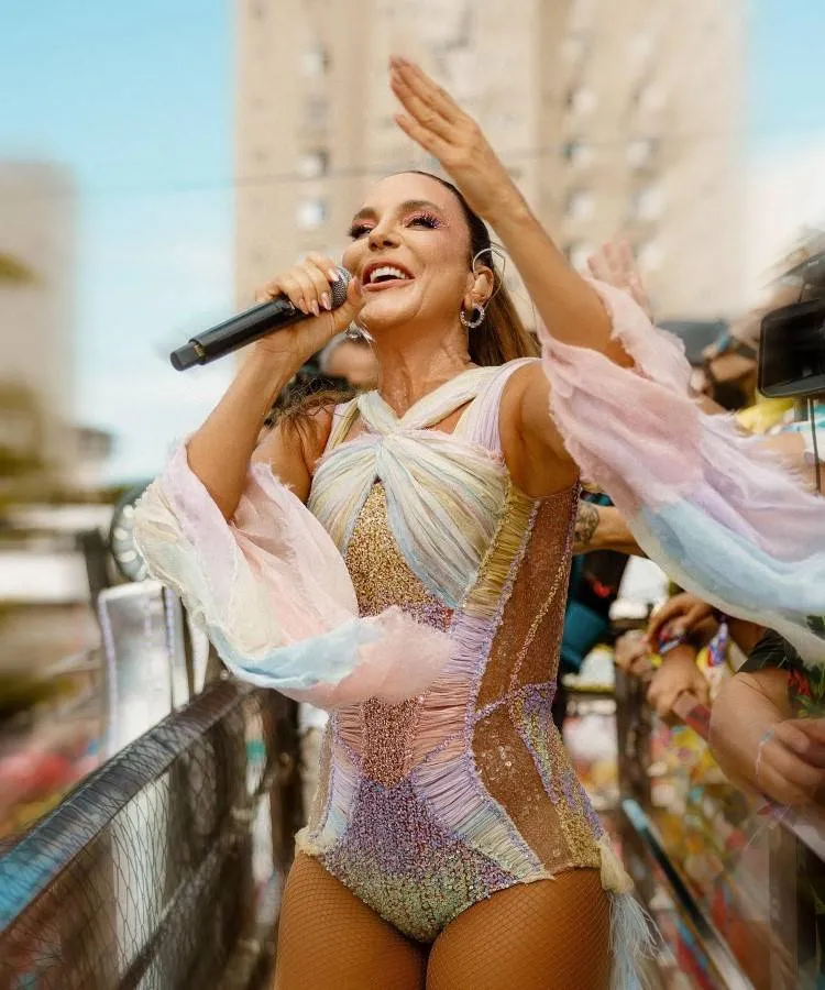 Ivete Sangalo vibrante em ação, possivelmente durante um desfile. Ela sorri animada, segurando um microfone, com uma mão levantada. Veste um traje colorido e brilhante. Ao fundo, prédios e uma multidão em um ambiente urbano e festivo.