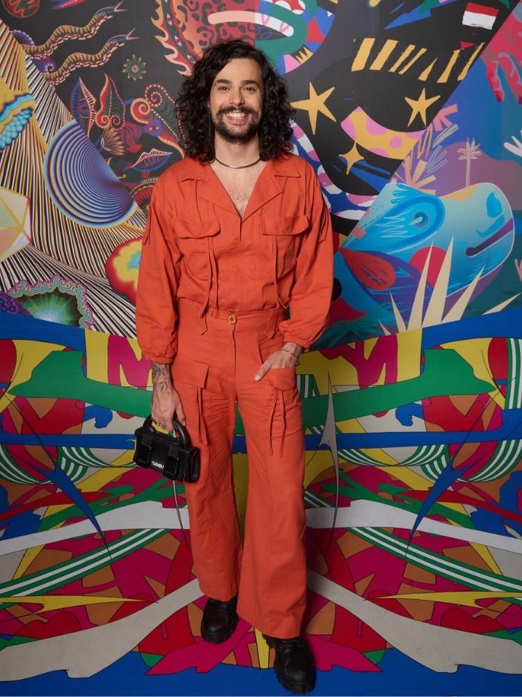 Vitor DiCastro de macacão laranja em frente a uma parede colorida 