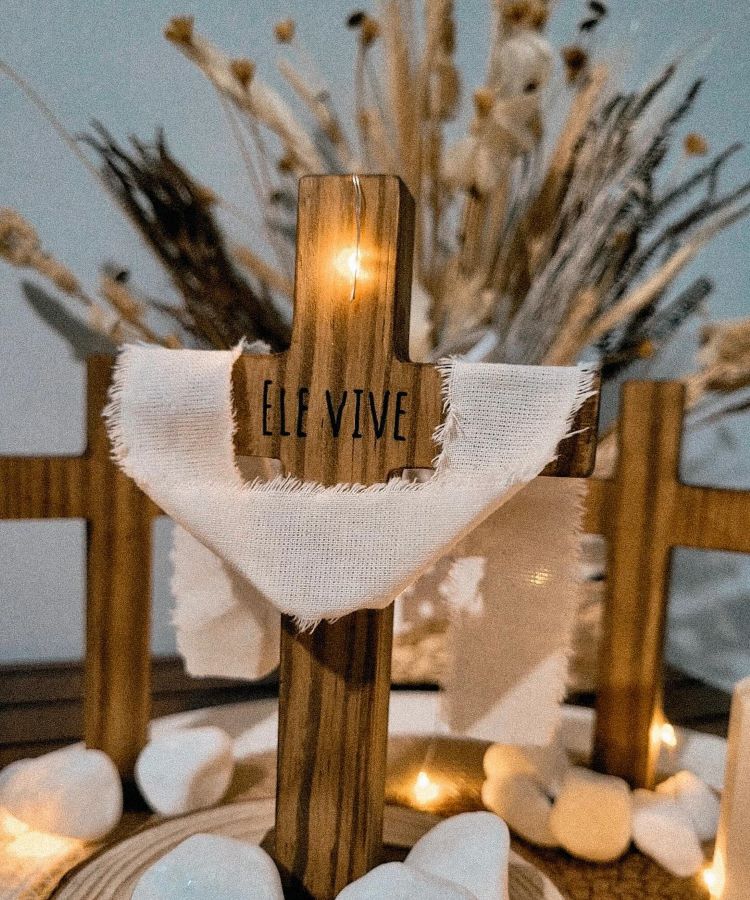 Cruz de madeira com a inscrição “Ele Vive”, envolvida por tecido branco e cercada de pedras decorativas brancas. Ao fundo, arranjo de flores secas em tonalidades neutras, trazendo um clima rústico e sereno, com iluminação suave.