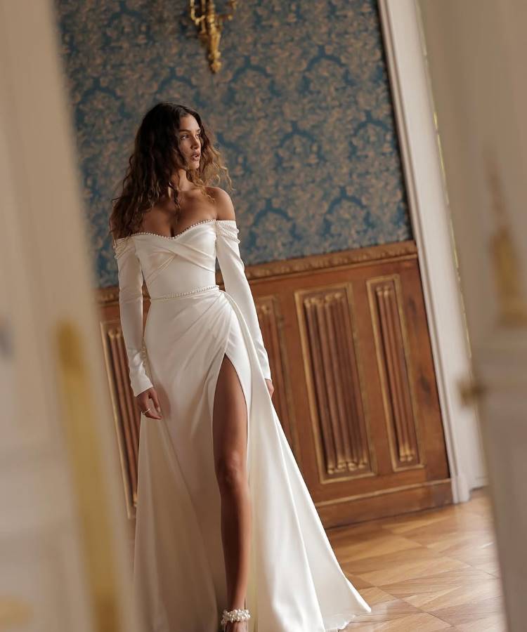 Noiva em vestido branco elegante, com decote ombro a ombro, mangas longas e fenda alta. O design sofisticado combina drapeado fluido e detalhes delicados, destacando feminilidade. Fundo clássico com papel de parede azul e molduras douradas complementa o visual refinado.





