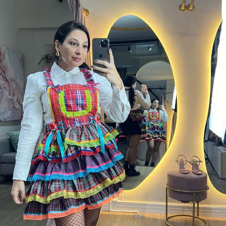 Mulher tira selfie no espelho, vestindo uma camisa branca e um vestido junino vibrante. O vestido é xadrez com camadas coloridas e fitas, exibindo um visual alegre e tradicional de festa junina.


Fontes





