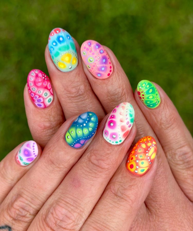 A imagem destaca unhas ovais decoradas com um design vibrante e psicodélico. Cada unha combina tons vivos de rosa, azul, verde, amarelo e laranja, formando padrões orgânicos e abstratos. Pequenos pontos brancos adicionam detalhes delicados e artísticos.
