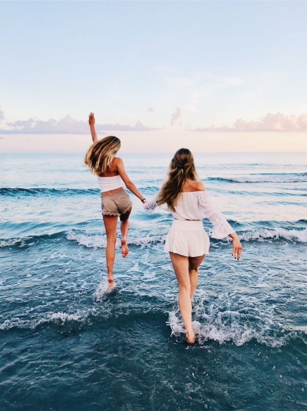Duas mulheres loiras de pele clara (uma usando top branco e shorts bege e a outra usando blusa branca e shorts branco) de mãos dadas entrando no mar