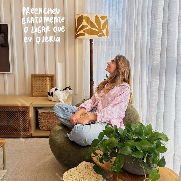 Mulher sentada em uma poltrona verde, usando camisa rosa e jeans, olha para cima sorrindo. Um gato repousa em um móvel próximo. A sala iluminada possui abajur estampado, plantas e cortinas claras, criando um ambiente acolhedor.





