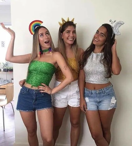 Foto de 3 mulheres de pele clara com cropped metalizados, shorts e uma usa tiara de arco-íris, outra de sol e outra de lua e estrelas