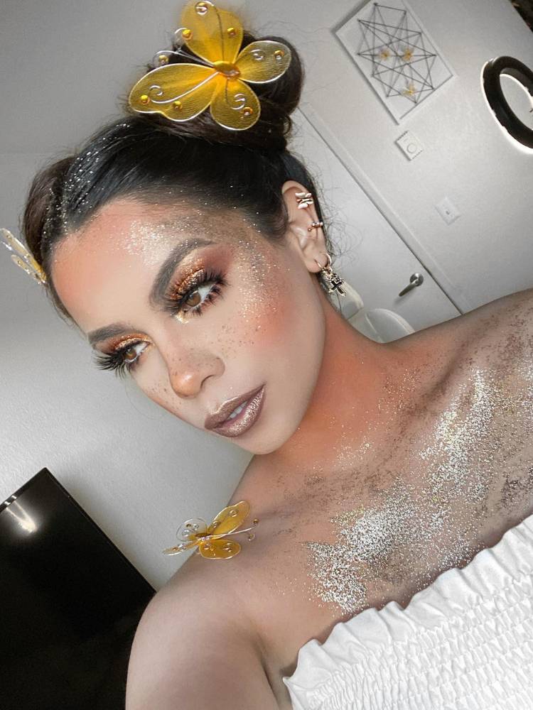 Mulher de pele clara e cabelo preto preso com enfeites de borboleta amarela, com o colo todo cheio de glitter prata com borboleta amarela e make glow. 