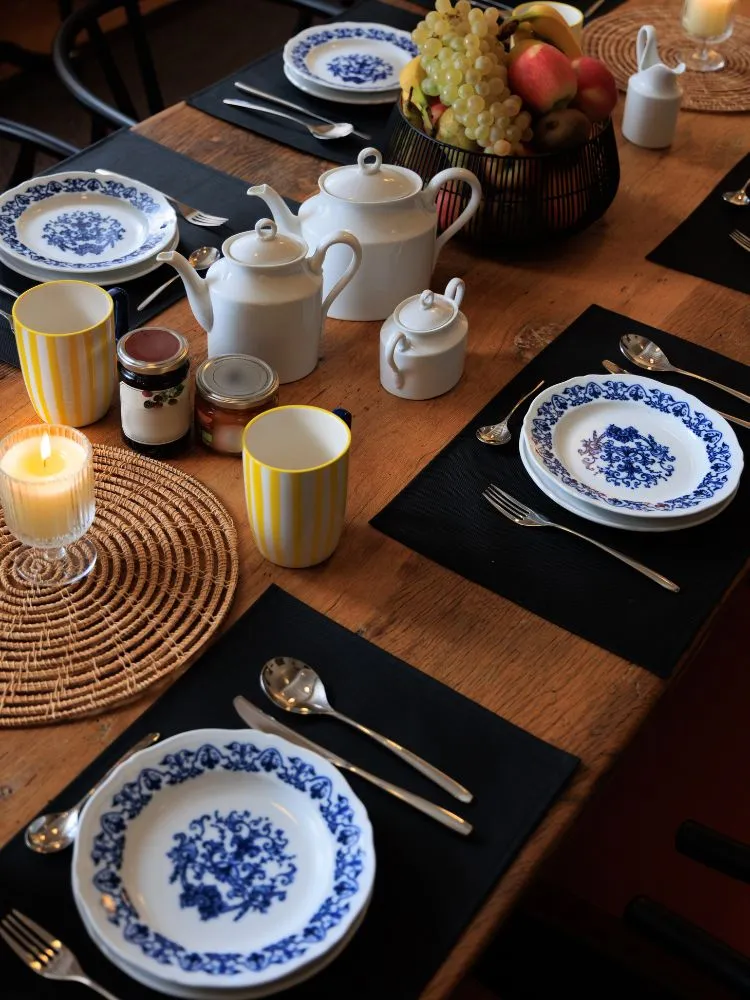 Mesa posta em estilo português com uma mesa de madeira com um pano preto cobrindo-a. Há quatro pratos brancos com desenhos florais azuis, um em cada canto da mesa. Cada prato tem uma colher de prata ao lado. Há também quatro jogos americanos de palha sob os pratos. No centro da mesa, há um cesto de frutas com uvas e maçãs. Há também um bule e um açucareiro brancos, e duas velas listradas de amarelo e branco. A imagem transmite uma sensação de aconchego e tradição.
