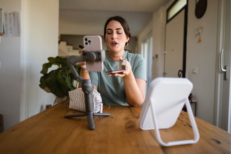 Uma mulher blogueira de maquiagem, com camiseta verde e olhos fechados, fala enquanto se grava com um celular em um tripé. Ela segura um produto de beleza, com um espelho de mesa à direita, criando conteúdo em casa.

