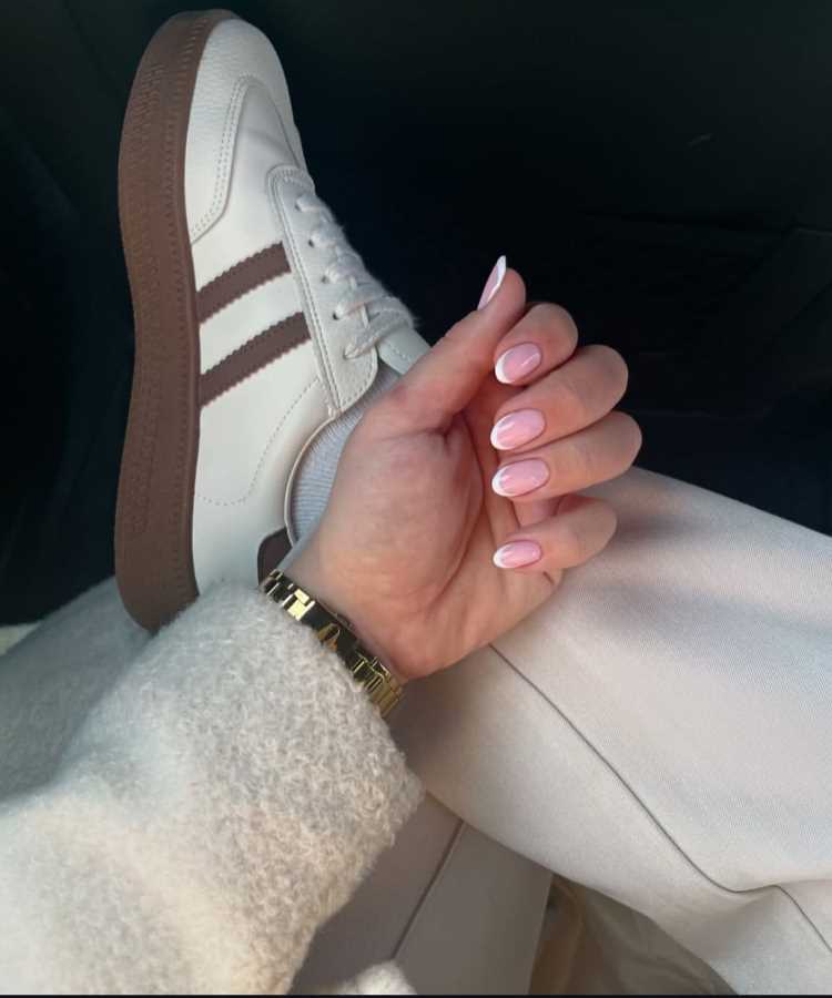 Mão feminina com unhas pintadas de esmalte rosa claro, segurando levemente a perna, vestindo calça bege e tênis branco com listras marrons. Detalhes incluem relógio dourado no pulso e casaco felpudo branco, compondo visual casual e elegante.