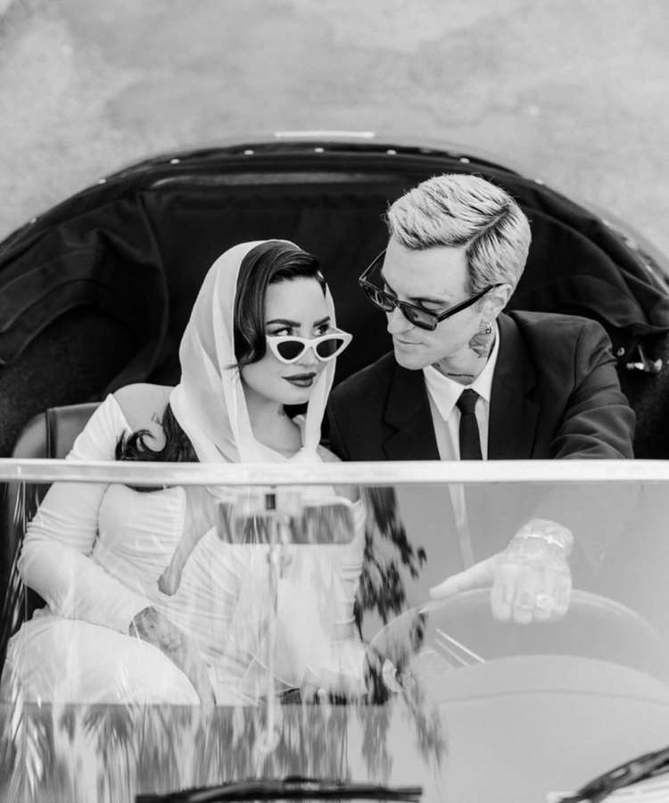: Em preto e branco, Demi Lovato e um homem estão sentados em um carro vintage. Ela, com um lenço na cabeça e óculos de sol, olha para ele, que está de terno e óculos, olhando para ela. A cena transmite um ar romântico.