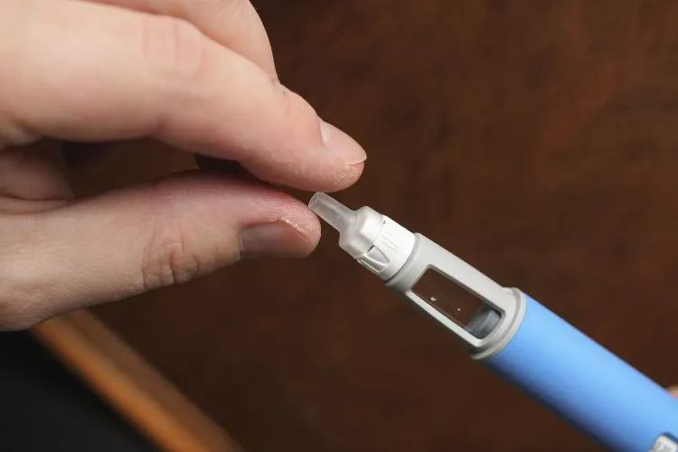 Close nas mãos de uma pessoa ajustando cuidadosamente uma ponta transparente em uma caneta injetora azul-clara, destacando o preparo do dispositivo médico antes da aplicação da injeção. Cenário enfatiza segurança e precisão.