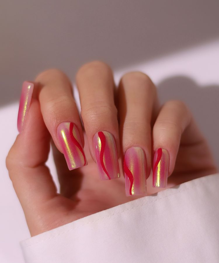 A imagem mostra unhas longas em formato quadrado, decoradas em tom rosa perolado com efeito furta-cor. Linhas onduladas em vermelho criam contraste moderno, conferindo um toque artístico e elegante ao design. Ideal para um visual sofisticado.