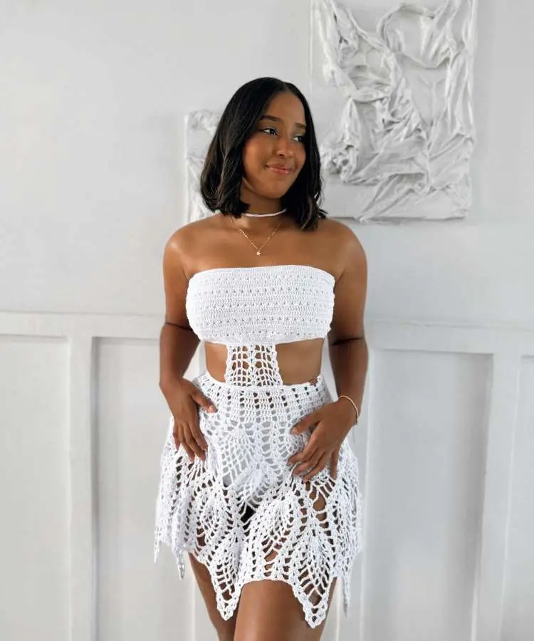 A mulher veste um conjunto branco elegante com detalhes vazados, posando em um ambiente minimalista. Ela segura um celular, refletindo-se no espelho ao lado de folhas decorativas.