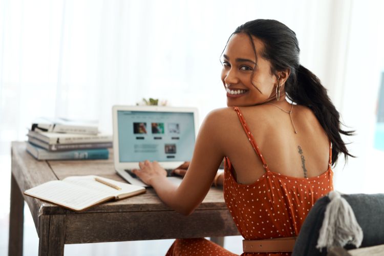 Mulher sorridente em mesa de madeira, usando laptop, caderno e livros ao redor. A cena reflete criação de conteúdo digital, ideal para representar a diferença entre influencer e blogueira, destacando trabalho criativo e engajamento em plataformas online.
