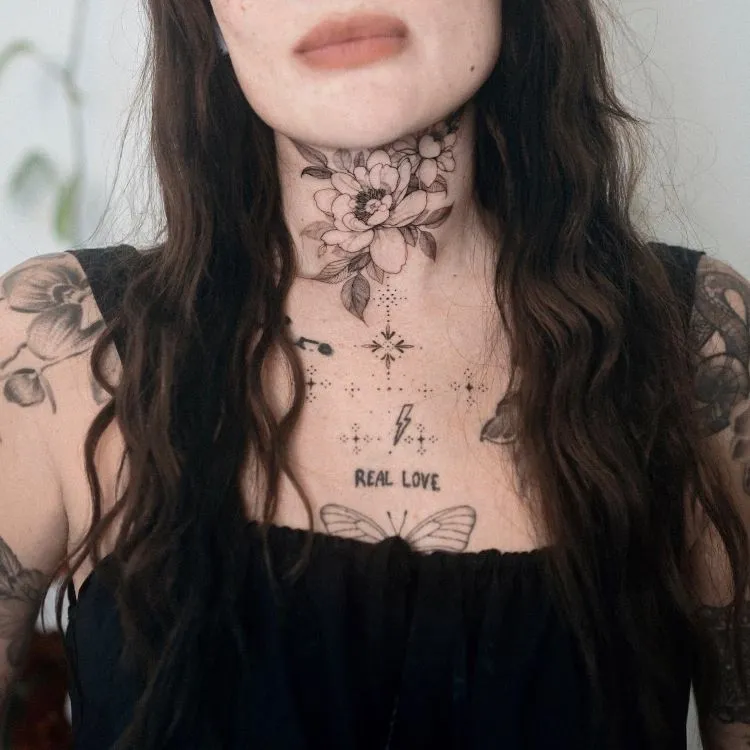 A imagem mostra uma pessoa com cabelo longo e ondulado. Ela tem uma tatuagem no pescoço de uma grande flor detalhada em preto e cinza, que se estende até o peito. A tatuagem inclui outros elementos como estrelas, um relâmpago, a frase "REAL LOVE" e uma borboleta.


