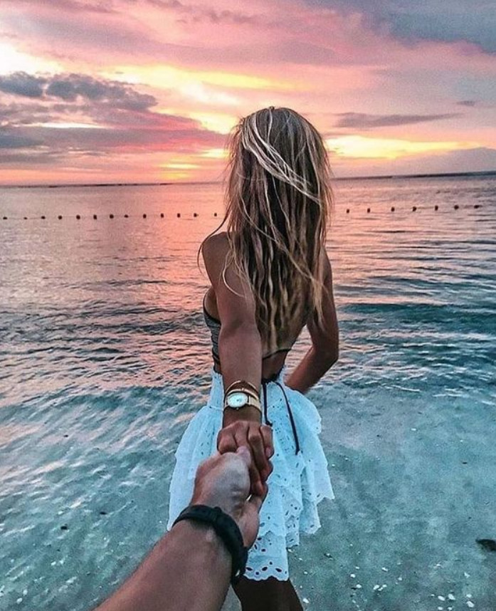 Mulher de pele clara usando top prateado e saia branca, de mãos dadas com homem de pele clara, na beira do mar ao pôr do sol