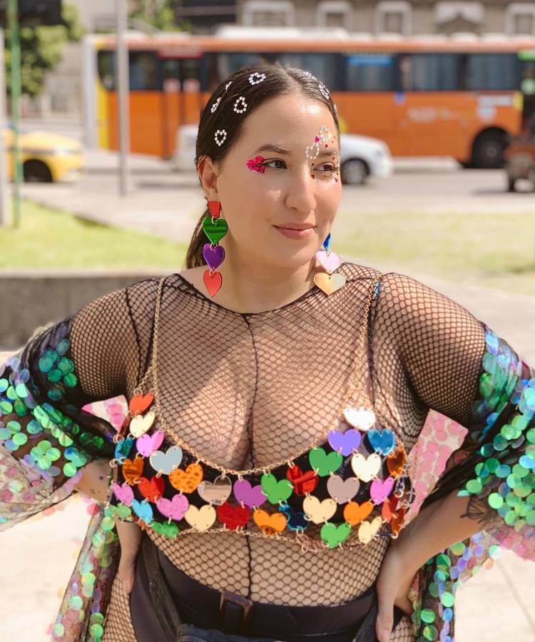 A mulher exibe um visual vibrante e carnavalesco, com maquiagem brilhante decorada por pedrinhas, penteado com flores aplicadas, brincos grandes de corações coloridos e um top de correntes com corações sobre uma blusa de tela preta, irradiando alegria.