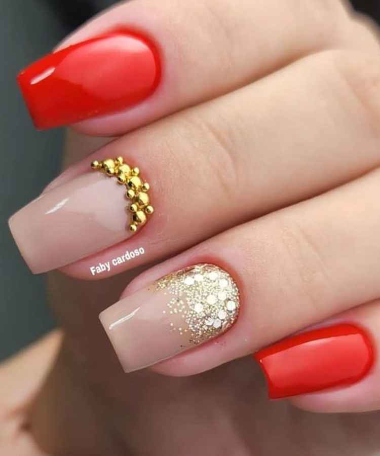 Pessoa de pele clara com unhas quadradas decoradas vermelhas com glitter dourado e pedrarias.