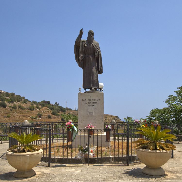 A imagem mostra a estátua de São Conrado em um pedestal, com a inscrição "A San Corrado, la Sua Città Devota". O santo, em postura serena, abençoa com uma mão levantada. Flores e plantas decoram o monumento.