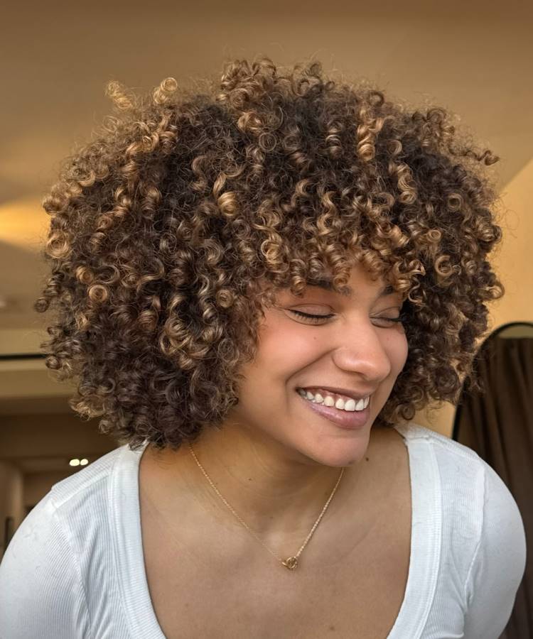 A imagem mostra uma mulher sorrindo com os olhos fechados, exibindo cabelos cacheados curtos e volumosos em tons de castanho e loiro. Ela veste uma blusa branca simples e um colar dourado delicado. A expressão dela é de alegria e relaxamento.







