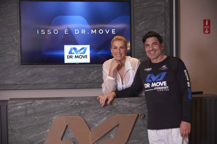 Um homem e uma mulher sorriem, apoiados em um balcão da clínica de longevidade DR MOVE. Ele veste uma camiseta preta da marca, e ela uma blusa branca. Acima deles, uma tela exibe a frase "ISSO É DR. MOVE".