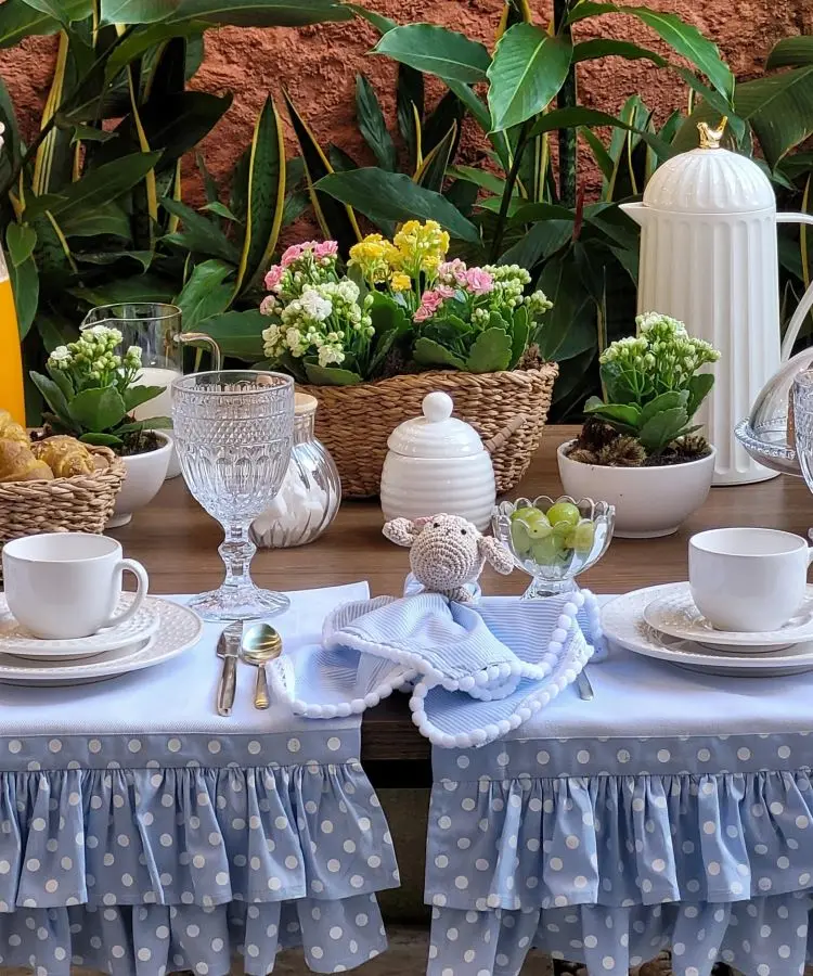 Mesa decorada com tema de café da manhã, com toalha azul de bolinhas brancas e babados, louças brancas, taças de vidro, enfeite de crochê de ovelha, arranjos de flores coloridas e plantas ao fundo, criando ambiente alegre e acolhedor.