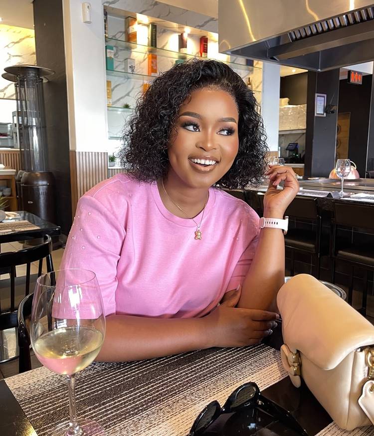 Mulher sorridente com cabelo cacheado curto, vestindo blusa rosa e relógio branco. Ela está em um restaurante moderno, com taça de vinho sobre a mesa. Ao fundo, prateleiras iluminadas com garrafas coloridas, criando um ambiente acolhedor e sofisticado.