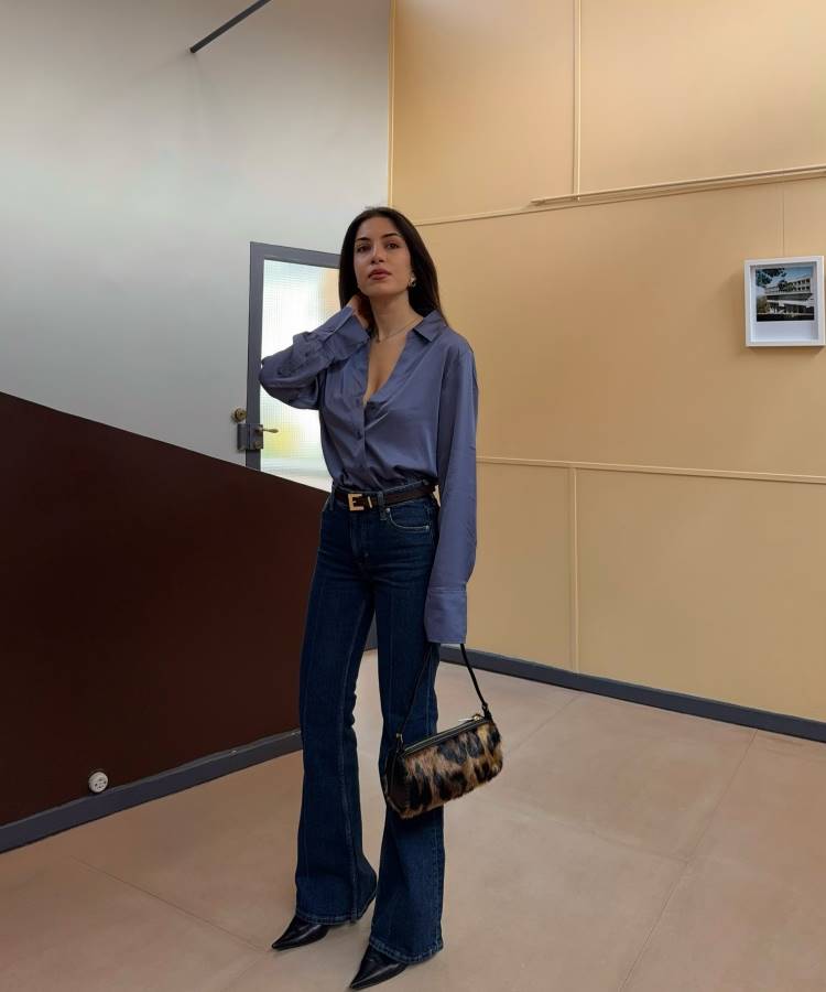Mulher posa em ambiente interno com camisa de cetim azul ajustada, jeans escuro de barra flare e cinto preto com fivela dourada. Completa o look com bolsa de ombro peluda e botas de couro. Inspiração moderna para looks de trabalho elegantes.





