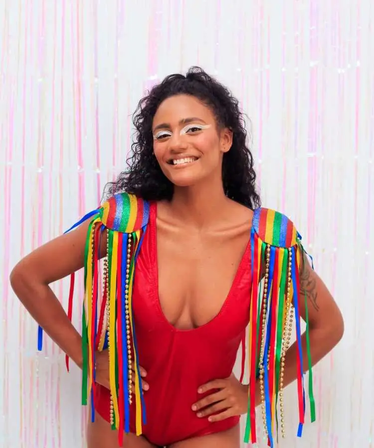 Uma mulher sorridente veste um body vermelho e ombreiras coloridas decoradas com fitas e correntes douradas. Seu cabelo cacheado está solto, e sua maquiagem branca destaca os olhos. O fundo é composto por tiras rosa e branco.