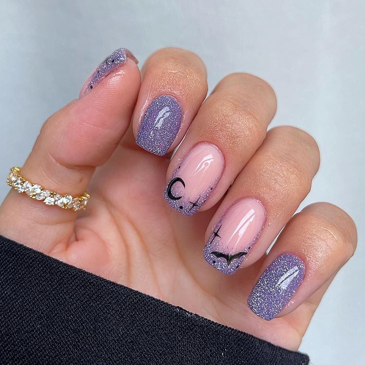 Mão com unhas médias quadradas decoradas noite de Halloween, com esmalte de glitter lilás e desenhos de lua, morcego e estrelas em preto