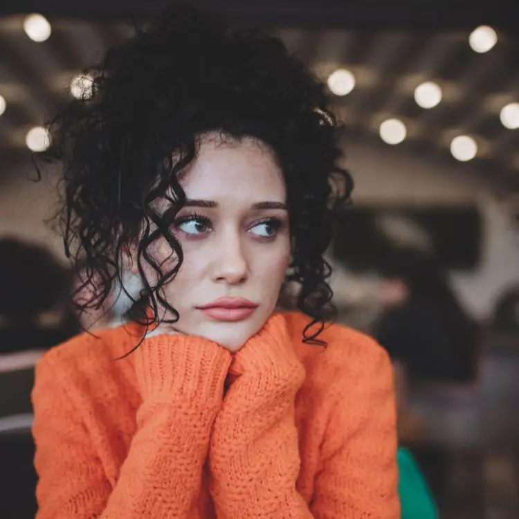 foto de mulher com braços apoiados na mesa e no rosto, veste blusa laranja