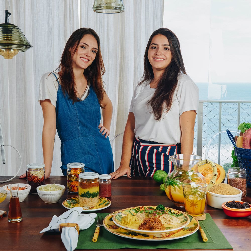 Isabelle Drummond ao lado de mulher de pele clara. Elas estão em frente a um balcão com comidas