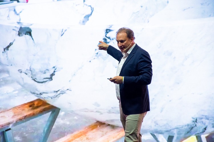 Alex Allard de terno azul, camisa clara e calça bege palestrando em evento