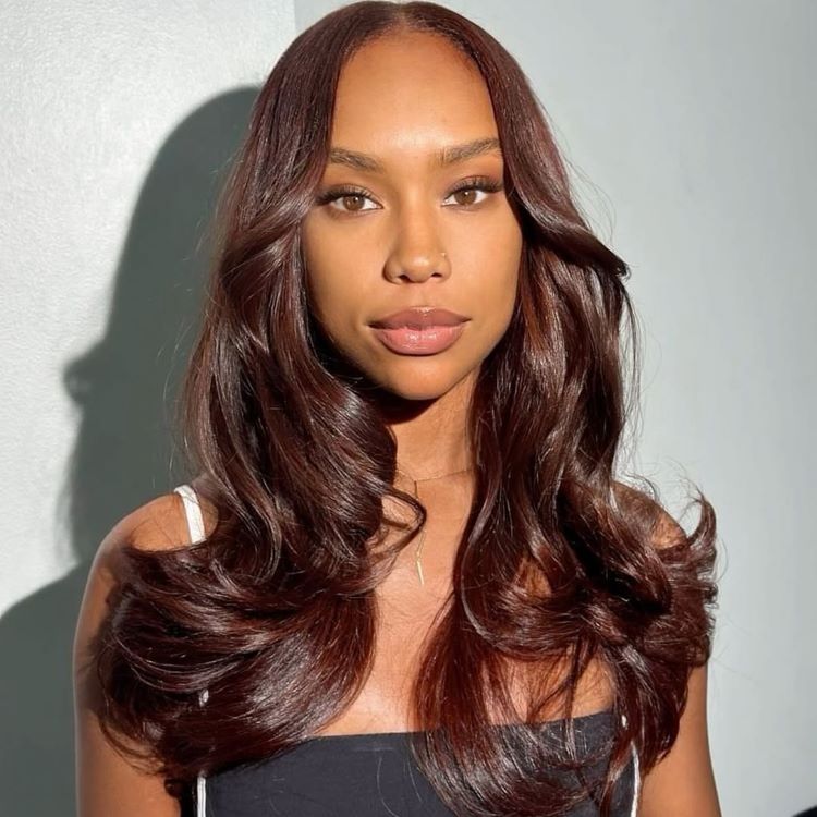 Mulher com cabelos longos e ondulados em tons de castanho avermelhado brilhante. Ela tem pele iluminada, maquiagem neutra com batom nude, veste uma regata branca e um top preto. Fundo minimalista em tom claro realça os detalhes da imagem.