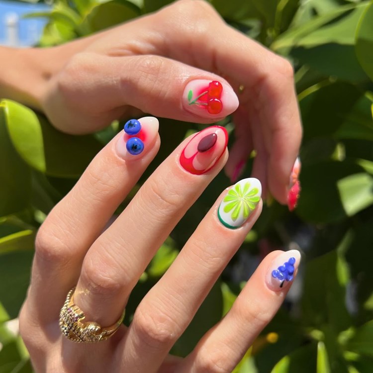 Unhas decoradas com desenhos de frutas coloridas, como cerejas, uvas, limão e mirtilos, sobre base clara. A mão usa um anel dourado brilhante. Fundo com folhas verdes iluminadas pelo sol, criando um cenário vibrante e natural.