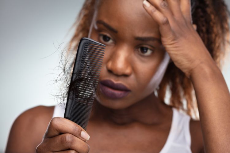 Mulher de pele negra usando regata branca e olhando para pente com muitos fios de cabelo com semblante de preocupada