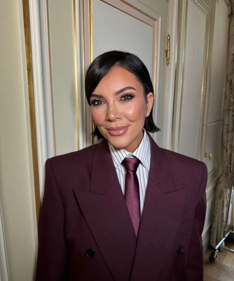 Kris Jenner posa sorridente, vestindo um terno vinho com camisa listrada e gravata. Ela tem cabelo curto e escuro, maquiagem destacando os olhos e um semblante elegante em um ambiente interno.