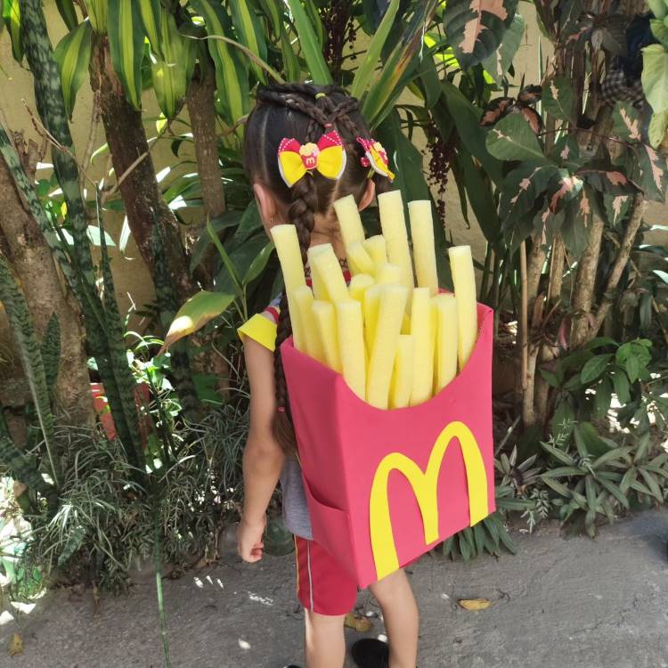 A imagem mostra uma criança usando uma mochila maluca em forma de caixa de batatas fritas do McDonald's. A mochila é vermelha com o icônico arco dourado e repleta de "batatas" amarelas. A criança, de costas, tem o cabelo preso em tranças com laços coloridos. Ela está em um ambiente com plantas verdes, possivelmente um jardim ou área externa.


