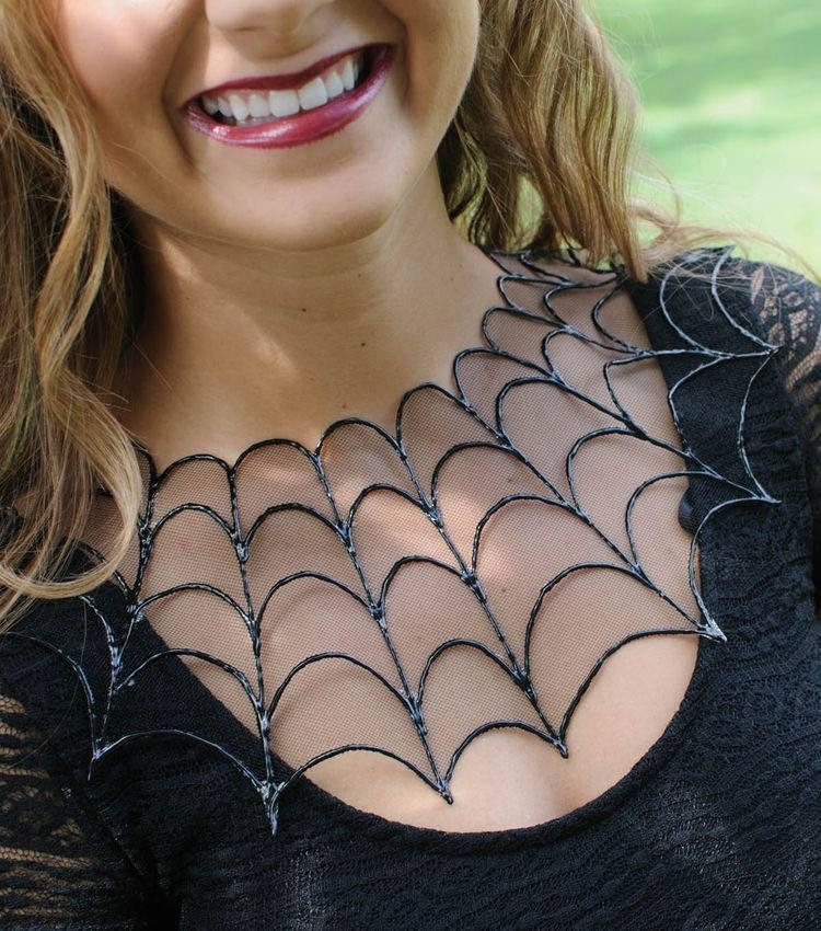 Zoom em colo de mulher de pele clara sorrindo, usando colar em forma de teia de aranha sobre roupa preta 
