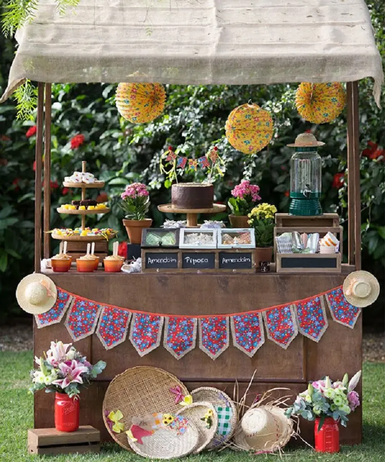 A imagem representa uma decoração de festa junina colorida ao ar livre. Contém mesa com teto repleta de comidas como pipoca, amendoim, doces, além de enfeites como bandeirinha, chapéus de palha, peneira, flores, etc