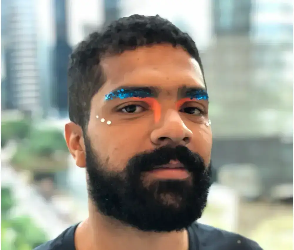 Maquiagem masculina para olho de carnaval com brilho e strass Foto de homem de pele morena com glitter flocado azul na sobrancelha, sombra laranja neon e strass