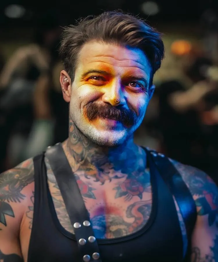Homem de pele clara tatuado usando sombra laranja no olho e espiral na ponta do nariz 