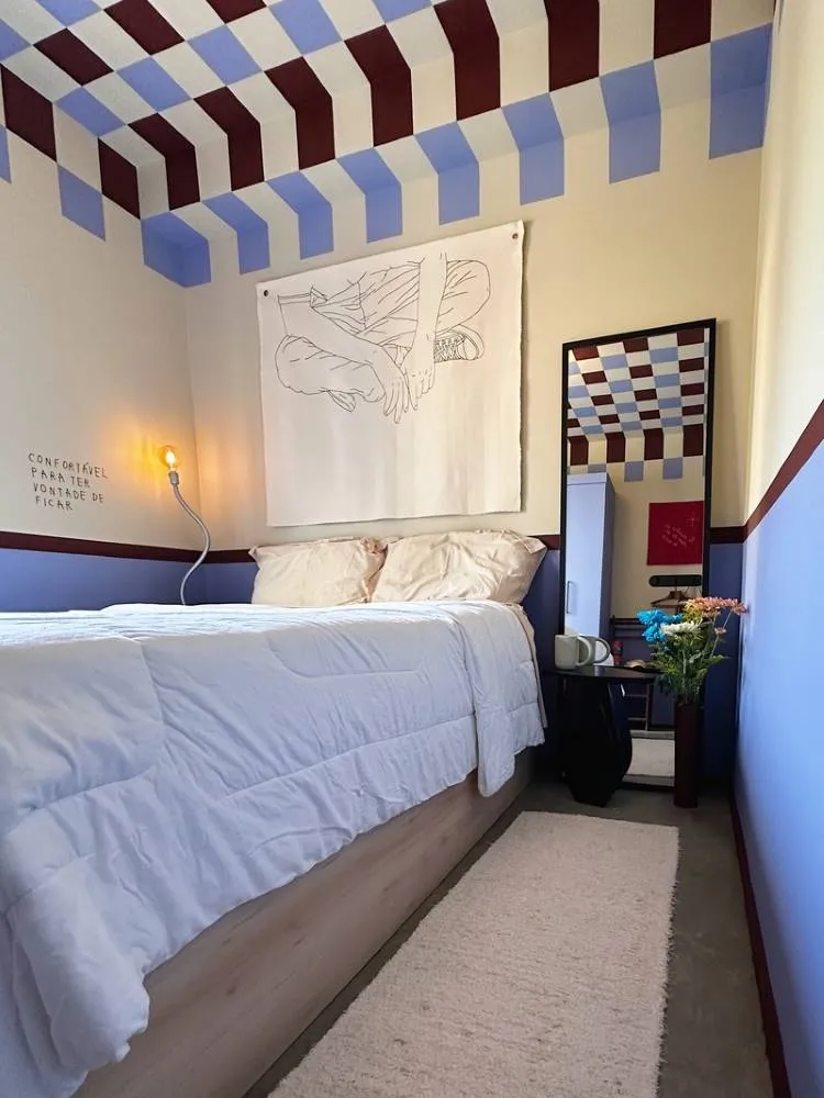 Esta imagem mostra um quarto com uma cama de casal e roupa de cama branca. O quarto possui um teto e paredes com um padrão geométrico em tons de azul e bordô. Um espelho alto reflete o padrão e há uma arte abstrata na parede.

