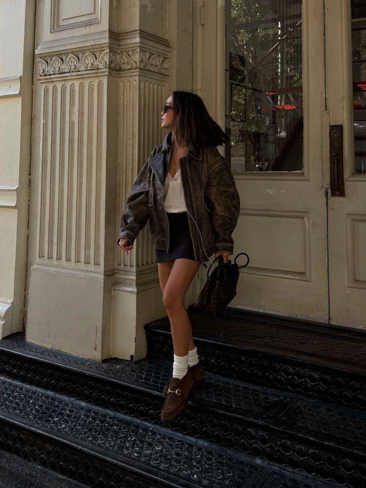Mulher de pele clara usando blusa branca, jaqueta biker de couro, meia 3/4, minissaia escura e mocassim de camurça marrom 