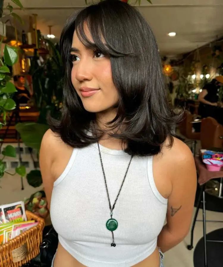 Mulher com cabelo médio castanho escuro, corte reto com pontas modeladas e franja cortina. Ela veste regata branca e colar com pingente verde. O ambiente interno possui plantas e decoração ao fundo, destacando o visual elegante.