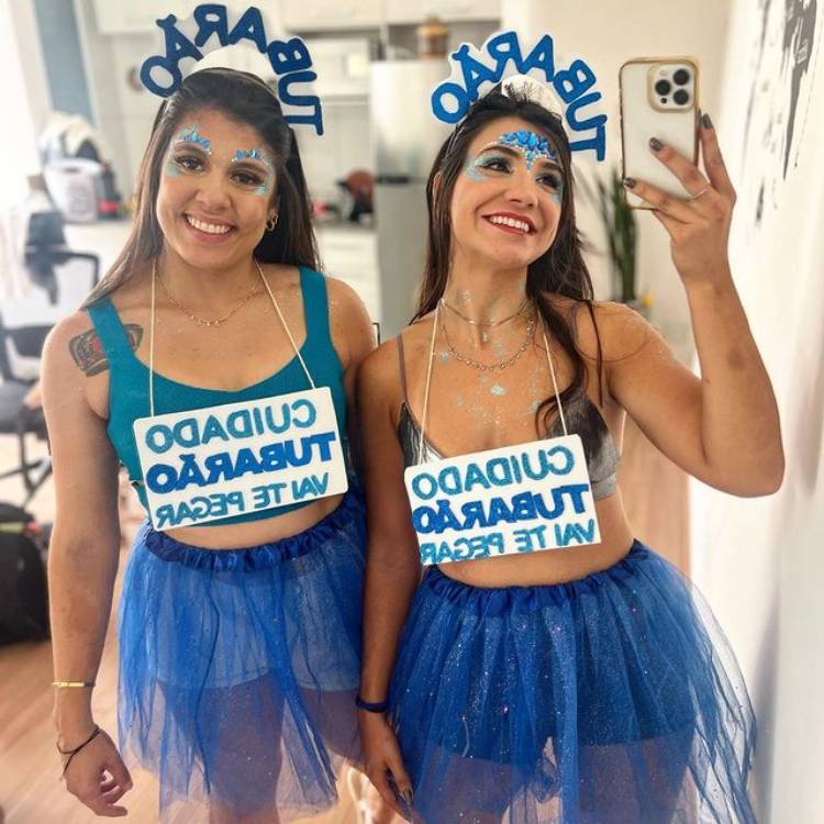Amigas em foto no espelho com top e saia azuis, tiara escrito tubarão e plaquinha com trecho "cuidado tubarão vai te pegar".